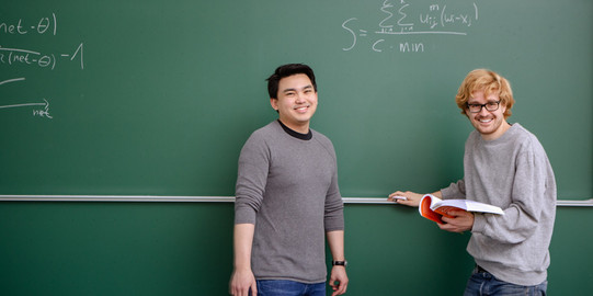 Zwei Studierende stehen vor der Tafel, lächeln in die Kamera