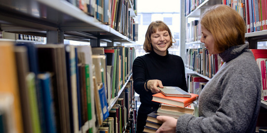 Two students stand between two bookshelves with books in hands in the library.